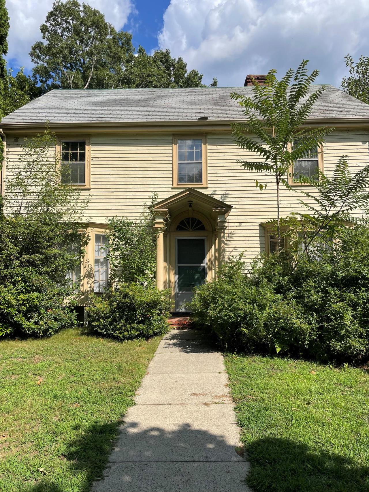 Image shows the Howard House, a yellow home with green shrubbery around the front. 